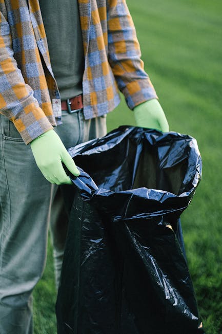 A person wearing a casual plaid shirt with yellow, grey, and blue checks, green gloves, and grey trousers standing outdoors on a grassy area, holding open a large black plastic garbage bag with a shiny finish. The person is preparing to gather and dispose of waste, with the bag's opening held wide to allow for easy filling. The background shows a lush green lawn, indicating the scene takes place in a garden or park setting. The image is relevant to rubbish removal services offered by Waste Disposal Balham, illustrating a private waste collection or on-site clearance process for garden or outdoor waste management. Natural daylight highlights the texture of the gloves and the glossy surface of the trash bag, emphasizing the activity of waste handling in an environmentally conscious manner.