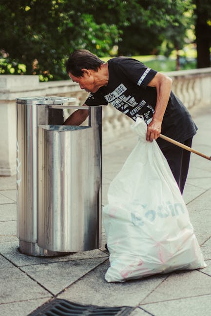 A person wearing a casual plaid shirt with yellow, grey, and blue checks, green gloves, and grey trousers standing outdoors on a grassy area, holding open a large black plastic garbage bag with a shiny finish. The person is preparing to gather and dispose of waste, with the bag's opening held wide to allow for easy filling. The background shows a lush green lawn, indicating the scene takes place in a garden or park setting. The image is relevant to rubbish removal services offered by Waste Disposal Balham, illustrating a private waste collection or on-site clearance process for garden or outdoor waste management. Natural daylight highlights the texture of the gloves and the glossy surface of the trash bag, emphasizing the activity of waste handling in an environmentally conscious manner.