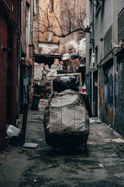 A narrow urban alleyway cluttered with a large, grey, industrial-style rubbish bag in the foreground, positioned on a rough asphalt surface that appears stained and uneven. Behind this bag, there are multiple piles of mixed waste materials, including cardboard boxes, plastic bags, and miscellaneous debris, stacked along a fence and against a brick wall with peeling paint. The alley is flanked by tall, weathered buildings with metal fire escapes and graffiti on the walls, suggesting a neglected, urban environment. Bare tree branches are visible above, indicating a colder season. The scene is illuminated by natural daylight, casting subtle shadows and highlighting the textures of the waste and surrounding surfaces. This setting reflects a typical private or independent rubbish collection scene, where waste is temporarily stored before professional clearance, aligning with services offered by Waste Disposal Balham for comprehensive rubbish removal and waste management solutions.