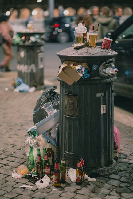 A black, cylindrical public trash bin with a domed, flat top located on a cobbled street. The top surface is cluttered with various discarded items, including two paper cups with remnants of whipped cream and straws, a crumpled paper napkin, and a plastic container. The front of the bin features a small, rectangular opening for waste disposal, with visible layers of dirt and grime. Surrounding the base, there are multiple empty glass bottles, some with labels partially intact, along with crumpled tissues and other small bits of litter scattered on the cobblestone ground. To the left side of the bin, a black garbage bag leans against it, containing additional rubbish, while on the right, a pink suitcase is partially visible. In the background, blurred figures of people and parked cars suggest an urban outdoor setting, with ambient lighting indicating either late afternoon or early evening. Waste Disposal Balham's services may be needed for alternative waste collection in such public spaces or private rubbish clearance scenarios, highlighting the importance of ongoing waste management efforts to maintain street cleanliness and hygiene.