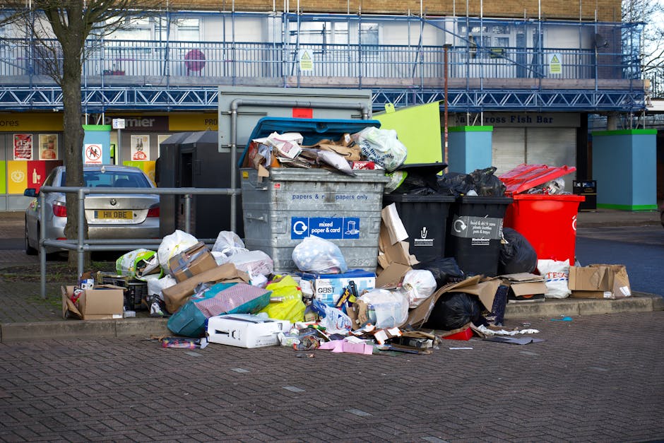 A narrow urban alleyway cluttered with a large, grey, industrial-style rubbish bag in the foreground, positioned on a rough asphalt surface that appears stained and uneven. Behind this bag, there are multiple piles of mixed waste materials, including cardboard boxes, plastic bags, and miscellaneous debris, stacked along a fence and against a brick wall with peeling paint. The alley is flanked by tall, weathered buildings with metal fire escapes and graffiti on the walls, suggesting a neglected, urban environment. Bare tree branches are visible above, indicating a colder season. The scene is illuminated by natural daylight, casting subtle shadows and highlighting the textures of the waste and surrounding surfaces. This setting reflects a typical private or independent rubbish collection scene, where waste is temporarily stored before professional clearance, aligning with services offered by Waste Disposal Balham for comprehensive rubbish removal and waste management solutions.