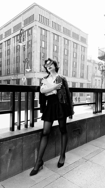 A black and white photograph of a young woman standing on a city sidewalk, in front of a modern multi-storey building with large rectangular windows and a geometric facade. She is dressed in a short skirt, high heels, dark tights, and a light-colored blouse, with a dark jacket draped over her arm. She is wearing glasses and has short, wavy hair, with a confident pose, one hand resting on her shoulder while holding the jacket. The background includes street elements such as utility poles and street lamps, and a few pedestrians can be seen in the distance, suggesting a busy urban environment. The scene captures a moment of fashion and city life, with the woman appearing poised against the backdrop of contemporary architecture and urban infrastructure, typical of a city setting relevant to private or independent waste collection in a busy urban context.