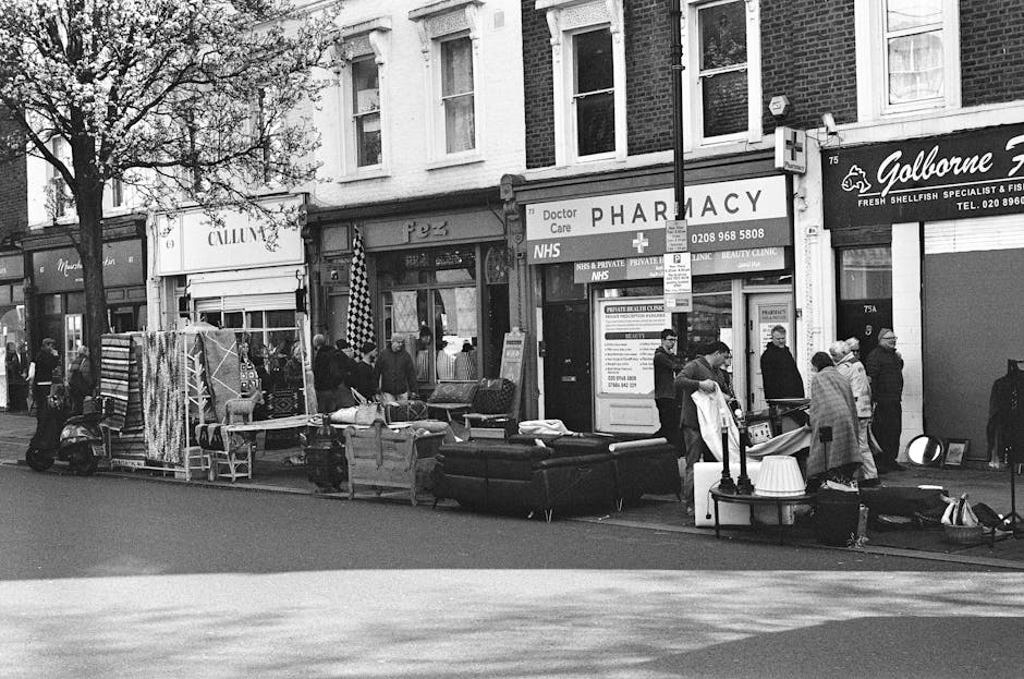 The image depicts a street scene featuring a row of small commercial establishments, including a pharmacy and a fish shop, with several pedestrians walking and standing along the pavement. In front of the storefronts, there is an outdoor display of various furniture items and household goods, such as sofas, chairs, and a table, arranged on the sidewalk. Some items appear to be upholstered or made from wood, with textures ranging from smooth to fabric-covered surfaces. A few people are engaged in examining or handling these objects, suggesting an informal sale or collection activity. To the left, a motorcycle is parked parallel to the curb, and additional smaller objects on the pavement include bags and possible miscellaneous goods. The background includes trees and building facades, with the scene captured under natural daylight, creating a typical urban environment where private collection or alternative waste handling may occur outside of official rubbish collection points. This setting relates to independent rubbish disposal efforts by local residents or businesses, consistent with private waste removal services such as those provided by Waste Disposal Balham.