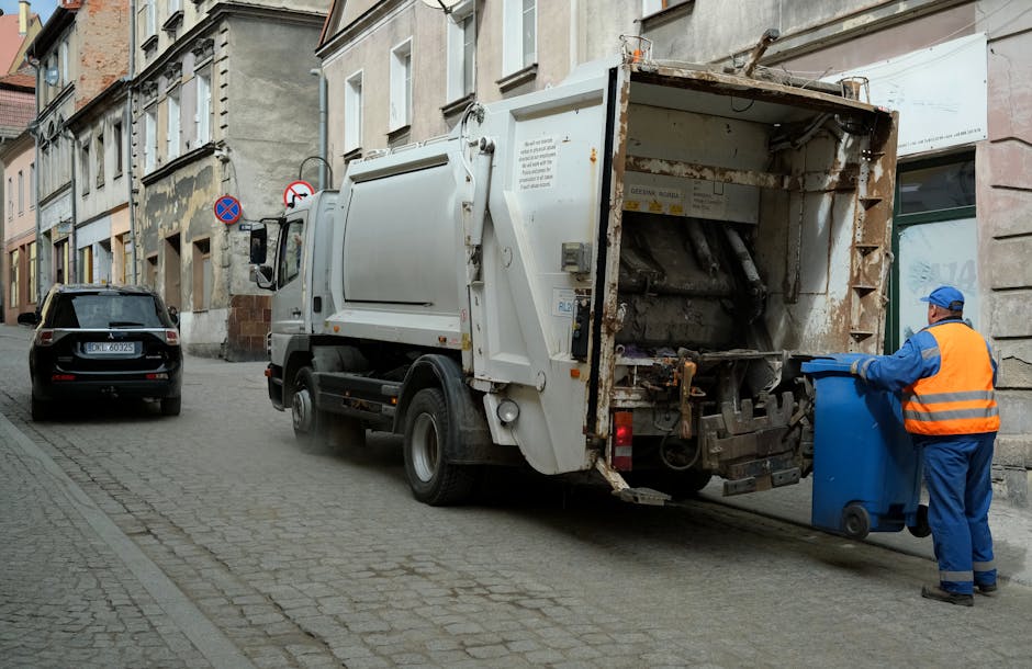 A municipal-style waste collection truck, primarily white with some rust and dirt on its rear loading area, is parked on a narrow cobblestone street in an urban setting. The rear compartment is open, revealing mechanical components and a collection area designed for compacting rubbish. A sanitation worker, dressed in a blue uniform and high-visibility orange safety vest, is placing a blue wheelie bin into the truck's receptacle. To the left of the truck, a black car is parked parallel to the curb. The surrounding environment includes older stone and brick buildings with multi-pane windows, some featuring weathered facades. Street signs indicating parking restrictions and no stopping are mounted on the building walls, contributing to the functional city street atmosphere. The scene is lit by natural daylight, with a subtle industrial tone emphasizing the routine nature of private waste collection by waste management services such as Waste Disposal Balham, highlighting an alternative approach to local authority rubbish collection.