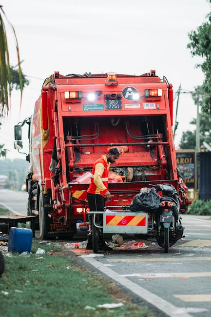 A worker dressed in a red and yellow uniform is performing rubbish collection on a street, standing next to the opening of a large red waste collection truck. The truck's back panel is lifted, revealing internal mechanisms for waste processing, and the worker appears to be loading or attending to waste materials. The scene includes a small black waste bag placed on a nearby trolley or platform, with some debris and discarded items scattered around the base of the truck. In the background, there are trees, power lines, and a roadside shop or building with signage, indicating an urban or suburban environment. The lighting suggests daytime with natural light, emphasizing the functional aspect of the waste collection process. This image aligns with private rubbish removal services, such as those offered by Waste Disposal Balham, providing on-site clearance of household or commercial waste materials in a professional manner without reliance on local authority collection.